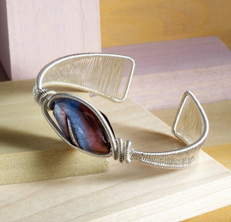 Puede incluir: Una pulsera de pu&ntilde;o de alambre plateado con una cuenta central de forma ovalada. La cuenta presenta una mezcla de colores azul y granate. El dise&ntilde;o de la pulsera incorpora un intrincado enrollado de alambre y detalles calados.