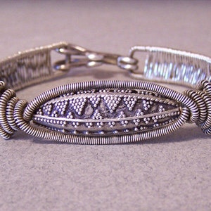 Puede incluir: Pulsera plateada con un centro ovalado con un patr&oacute;n detallado de puntos y tri&aacute;ngulos. La pulsera est&aacute; elaborada con un intrincado alambre y un cierre seguro. Una pieza de joyer&iacute;a hecha a mano.