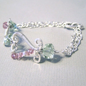 Op de afbeelding: Zilverkleurige armband met een hartvormig draadontwerp, versierd met roze en groene edelstenen. De armband heeft een sluiting met schakels en staat tegen een lichtgrijze achtergrond.