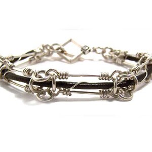 May include: A silver-toned bracelet with a black leather band. The bracelet features a series of interconnected silver wire loops and bars, creating a unique, handcrafted design. The clasp is a square shape. The bracelet is displayed against a white background.
