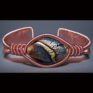 Puede incluir: Brazalete de pu&ntilde;o de cobre con una piedra de vidrio dicroico en tonos dorados y azules. La piedra est&aacute; engastada en un dise&ntilde;o envuelto en alambre, con el alambre de cobre formando la banda del brazalete. Una joya artesanal.