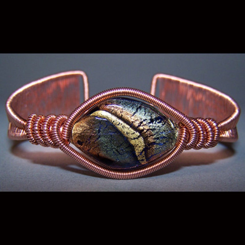 Puede incluir: Brazalete de pu&ntilde;o de cobre con una piedra de vidrio dicroico en tonos dorados y azules. La piedra est&aacute; engastada en un dise&ntilde;o envuelto en alambre, con el alambre de cobre formando la banda del brazalete. Una joya artesanal.