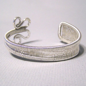Op de afbeelding: Een zilverkleurige manchetarmband met een geweven draadontwerp. De armband heeft een decoratief krul-element aan &eacute;&eacute;n uiteinde en een open ontwerp voor gemakkelijk dragen. De manchet is gemaakt van dunne, verweven draden, waardoor een textuur ontstaat.
