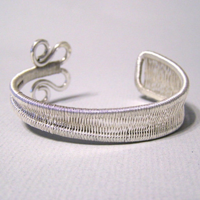 Op de afbeelding: Een zilverkleurige manchetarmband met een geweven draadontwerp. De armband heeft een decoratief krul-element aan &eacute;&eacute;n uiteinde en een open ontwerp voor gemakkelijk dragen. De manchet is gemaakt van dunne, verweven draden, waardoor een textuur ontstaat.