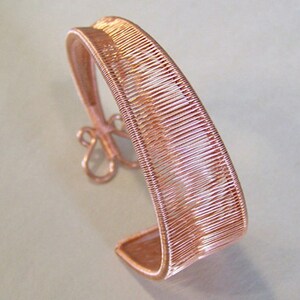 May include: A copper-coloured wire cuff bracelet. The bracelet has a woven design on the main band and a decorative floral element. The open cuff design allows for easy wear. The bracelet is a unique piece of jewellery.