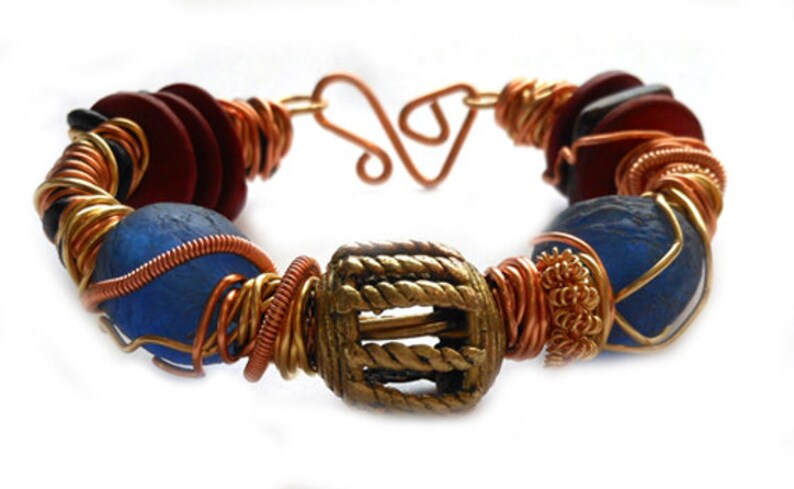 Pode incluir: Pulseira artesanal com uma mistura de materiais. Apresenta contas azuis e vermelhas, enroladas com fio de cobre e dourado. Uma conta central dourada em forma de barril adiciona um toque &uacute;nico. O fecho &eacute; um fecho de gancho e olho de cobre.