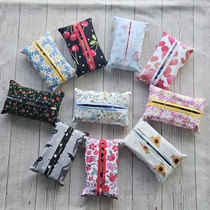 May include: A collection of rectangular fabric tissue holders in various patterns. Designs include daisies, cherries, hearts, sunflowers, and floral prints. Each holder has a zippered opening and is made of colorful fabric.