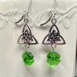 May include: A pair of silver-toned Celtic knot earrings with green four-leaf clover charms. The earrings have a triangular design with an interwoven knot pattern and a hook closure. The green charms are faceted and add a pop of color.