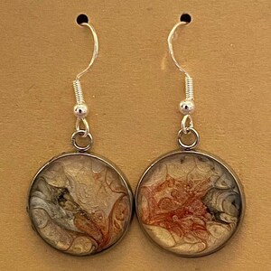 May include: A pair of silver dangle earrings with round, brown resin pendants. The pendants feature abstract, swirling patterns in shades of brown, orange, and white.