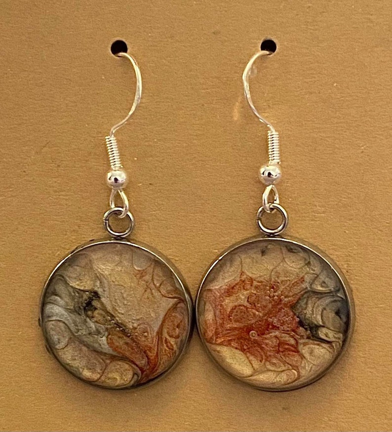 May include: A pair of silver dangle earrings with round, brown resin pendants. The pendants feature abstract, swirling patterns in shades of brown, orange, and white.