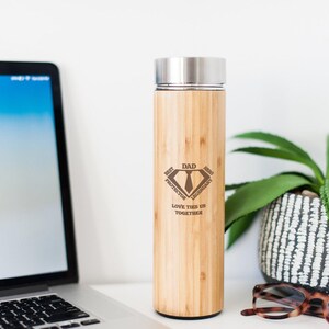 May include: A bamboo water bottle with a stainless steel lid. The bottle has a laser-etched design that reads "DAD BEST PROTECTOR LEGENDARY LOVE TIES US TOGETHER". A laptop, eyeglasses, and a potted plant are in the background.