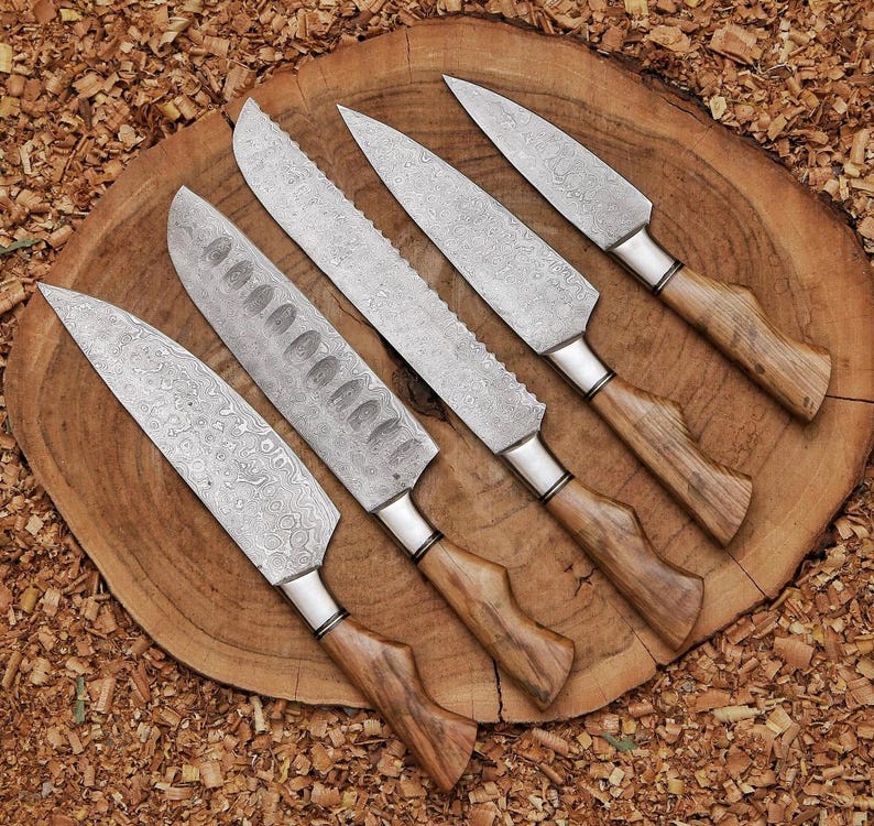 Peut inclure: Un ensemble de cinq couteaux de cuisine dot&eacute;s de lames en acier damas aux motifs ondul&eacute;s complexes et de manches en bois clair. Le set comprend un couteau de chef, un couteau &agrave; pain dentel&eacute; et trois couteaux utilitaires, dispos&eacute;s en diagonale.
