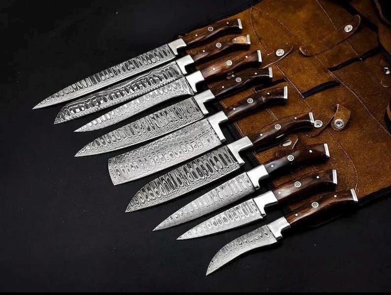 Peut inclure: Un ensemble de huit couteaux de cuisine avec des lames en acier damass&eacute; au motif ondul&eacute; et des manches en bois brun fonc&eacute; avec des rivets argent&eacute;s, dispos&eacute;s en diagonale sur un &eacute;tui en cuir brun avec des fermetures &agrave; pression et des coutures.