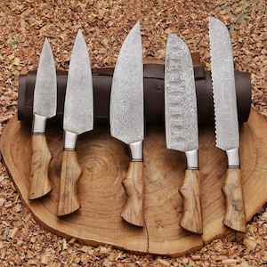Peut inclure: Un ensemble de cinq couteaux de cuisine avec des lames en acier damass&eacute; &agrave; motifs et des manches en bois brun clair. Les couteaux sont dispos&eacute;s en rang&eacute;e, pr&eacute;sentant diverses formes de lames dont une dentel&eacute;e, sur un &eacute;tui en cuir brun.