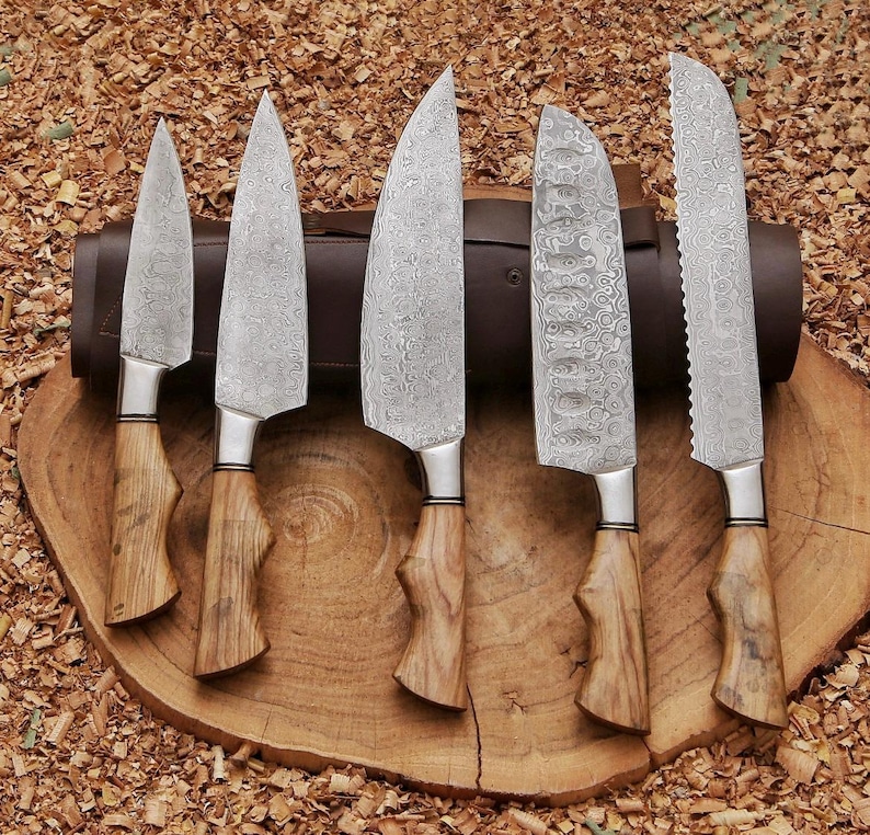 Peut inclure: Un ensemble de cinq couteaux de cuisine avec des lames en acier damass&eacute; &agrave; motifs et des manches en bois brun clair. Les couteaux sont dispos&eacute;s en rang&eacute;e, pr&eacute;sentant diverses formes de lames dont une dentel&eacute;e, sur un &eacute;tui en cuir brun.