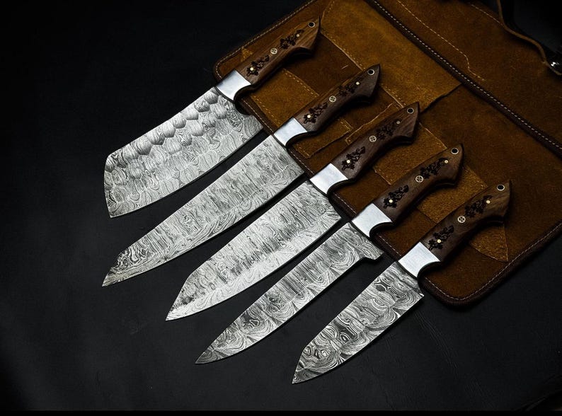 Peut inclure: Un ensemble de cinq couteaux de cuisine avec des lames en acier Damas pr&eacute;sentant un motif ondul&eacute; en couches. Chaque couteau poss&egrave;de un manche en bois brun fonc&eacute; avec des incrustations m&eacute;talliques florales et des rivets en laiton.