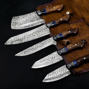 Peut inclure: Un ensemble de cinq couteaux dot&eacute;s de lames en acier damas &agrave; motifs et de manches en bois avec des accents bleus. Les couteaux sont dispos&eacute;s en rang&eacute;e, chacun avec une forme de lame distincte, incluant un couperet, un couteau de chef et des couteaux utilitaires.