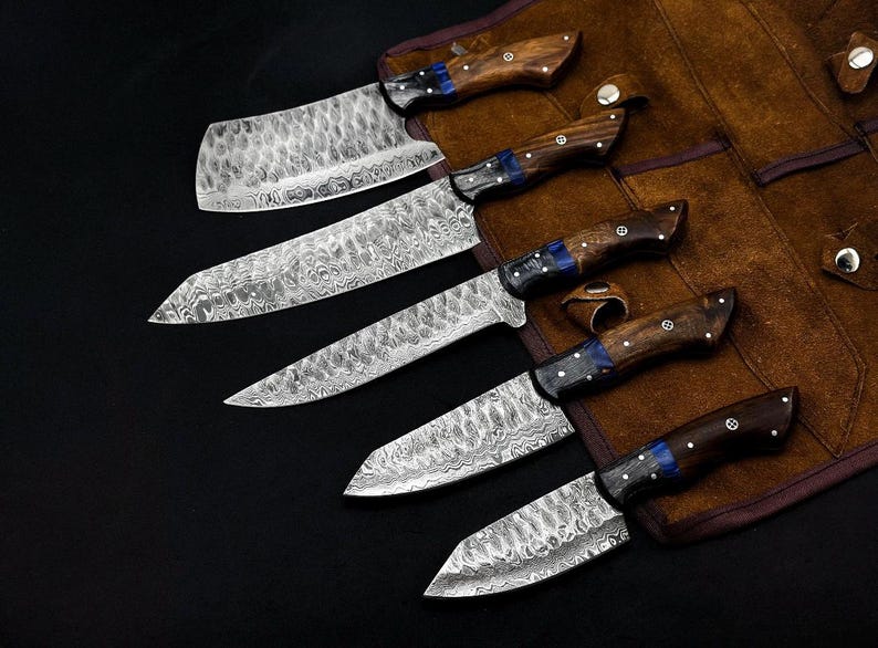 Peut inclure: Un ensemble de cinq couteaux dot&eacute;s de lames en acier damas &agrave; motifs et de manches en bois avec des accents bleus. Les couteaux sont dispos&eacute;s en rang&eacute;e, chacun avec une forme de lame distincte, incluant un couperet, un couteau de chef et des couteaux utilitaires.