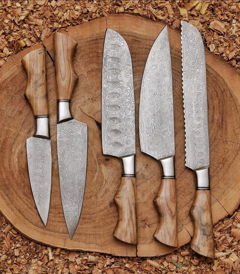 Peut inclure: Un ensemble de cinq couteaux de cuisine dot&eacute;s de lames en acier damas &agrave; motifs et de manches en bois brun clair. Les couteaux pr&eacute;sentent des formes de lame vari&eacute;es, incluant des bords pointus, incurv&eacute;s et dentel&eacute;s, avec une texture m&eacute;tallique ondul&eacute;e.