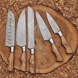 Peut inclure: Un ensemble de cinq couteaux de cuisine avec des lames en acier damass&eacute; &agrave; motifs et des manches en bois brun clair. Les couteaux varient en forme de lame, incluant des styles santoku, chef et office, avec une texture m&eacute;tallique ondul&eacute;e.