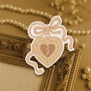May include: A heart-shaped sticker with a floral design and the letter "J" inside. The sticker has a ribbon and pearl chain design. The sticker is tan and brown, with a gold-colored frame in the background.