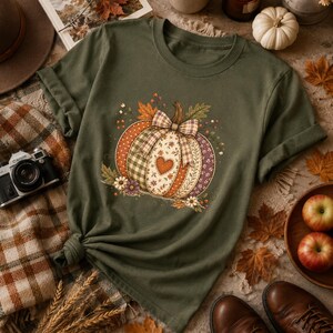 Op de afbeelding: Een olijfgroen T-shirt met korte mouwen en een centrale afbeelding van een pompoen, samengesteld uit verschillende stoffen met patronen zoals ruiten, bloemen en stippen, versierd met een grote geruite strik en een klein hartvormig detail.