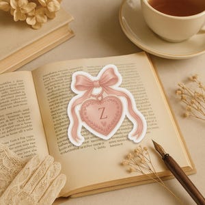 May include: A pink heart-shaped sticker with the letter "Z" inside, adorned with a bow and ribbons. The sticker is placed on an open book with a cup of tea in the background. The sticker is a decorative item.