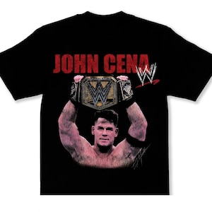 May include: Black t-shirt featuring a graphic of John Cena holding a championship belt. The text "JOHN CENA" is printed in red above the image, with the WWE logo in the upper right corner.