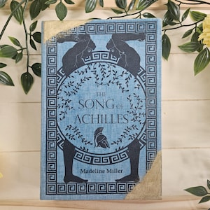 Puede incluir: Un libro de tapa dura titulado "The Song of Achilles" de Madeline Miller. El libro tiene una cubierta azul con ilustraciones negras de figuras y patrones de llaves griegas. Las esquinas están reforzadas con un material dorado. El libro está rodeado de vegetación y flores amarillas.