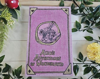 May include: A pink book cover with the title "Alice's Adventures in Wonderland" in a decorative font. The cover features an illustration of a rabbit in a circle and corner designs. The book is surrounded by greenery and a yellow flower.