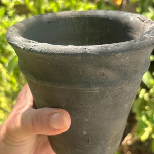 May include: A dark gray, weathered terracotta flower pot with a flared rim. The pot has a textured surface and is held in a hand against a backdrop of green foliage. Ideal for planting flowers or herbs.