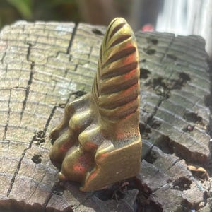 May include: A small, brass-colored decorative object with a textured, leaf-like design. The object has a rectangular base and a curved, ridged top, suggesting a stylized botanical form. The piece is set against a weathered wooden surface.