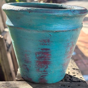 May include: A turquoise-colored, distressed-finish ceramic planter. The pot has a wide opening and a slightly flared rim. The turquoise paint is layered over a reddish-brown base, creating a weathered appearance. Ideal for indoor or outdoor use.