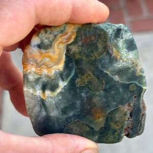 May include: A polished, irregular stone featuring a deep forest green base color with prominent, swirling bands of golden yellow and cream. The surface displays a smooth, mottled texture with dark green patches and subtle reddish-brown mineral inclusions.