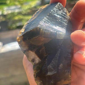May include: A raw, dark brown and black agate stone with a translucent, glassy texture and visible banding. The mineral specimen features sharp, angular edges and a polished, reflective surface that catches the sunlight, revealing deep, earthy tones.