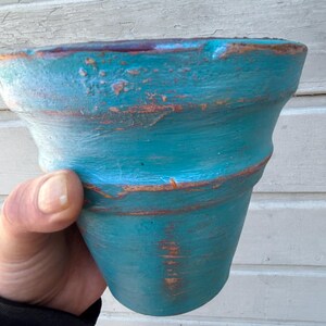 May include: A turquoise-colored, distressed ceramic flower pot. The pot has a wide, slightly flared rim and a textured surface with visible brushstrokes. The pot is held in a hand, showcasing its size and shape.