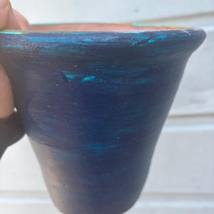 May include: A hand-painted, small, dark blue ceramic pot with teal and turquoise accents. The rim is a lighter shade of blue with hints of pink and yellow. The pot has a slightly flared shape, suitable for small plants or decorative use.