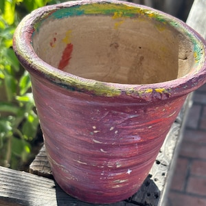 May include: A hand-painted, cylindrical terracotta flower pot. The pot is primarily a deep burgundy color with streaks of lighter red and white. The rim is painted with a band of green, yellow, and purple.