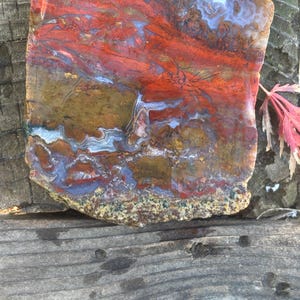 May include: A polished, rectangular stone with a vibrant display of colors. The surface features streaks of red, brown, blue, and white, creating a unique pattern. The stone is set against a weathered wooden background.