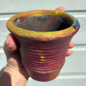 May include: A small, hand-painted terracotta pot with a ribbed exterior. The rim is painted with a mix of colors including yellow, orange, and blue. The pot is held in a hand.
