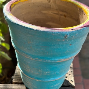 May include: A hand-painted, turquoise ceramic flower pot with a textured surface. The rim is accented with yellow, purple, and pink paint. The pot has a cylindrical shape with horizontal ridges.