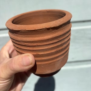 May include: A small, unglazed terracotta pot with a ribbed exterior. The pot is a warm reddish-brown color and has a rounded shape with a slightly flared rim. The pot is held in a hand.