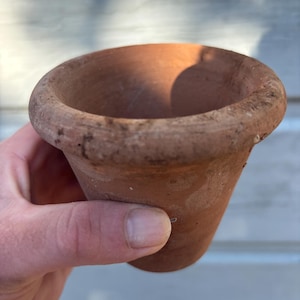 May include: A small, terracotta-colored clay pot with a rounded rim. The pot is held in a hand, showcasing its weathered texture and simple design. Ideal for small plants or succulents.