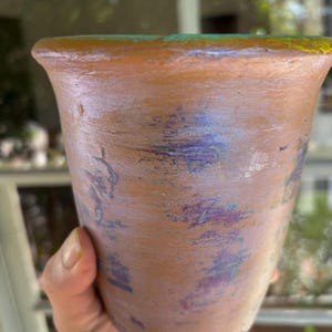 May include: A handmade, terracotta-colored ceramic pot with a flared rim. The pot features subtle purple and blue accents, adding a touch of color to the earthy tones. The rim is highlighted with green and yellow paint.