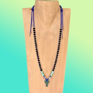 May include: A beaded necklace with black beads and a green pendant. The necklace has a blue cord and is displayed on a wooden stand. The pendant is a stylized cross shape. The necklace also features turquoise and white beads.