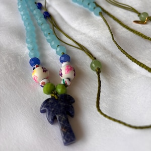 Puede incluir: Un collar hecho a mano con cuentas facetadas de color azul claro, detalles en azul oscuro y un colgante de palmera. El collar incluye cuentas decorativas con motivos florales y detalles de cordón verde. El colgante es de color azul oscuro.