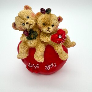 May include: A figurine of two teddy bears sitting on a red heart. The bears are light brown with dark eyes and small rose accents. One bear holds a red heart with the word "Love" and a rhinestone. The heart-shaped base reads "Love You."
