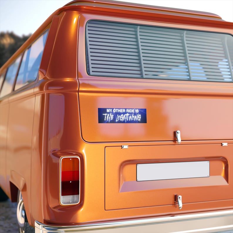 May include: Orange vintage van with a bumper sticker that reads "MY OTHER RIDE IS THE LIGHTNING" in white and blue text. The van has a rear window with horizontal blinds and a rectangular taillight.