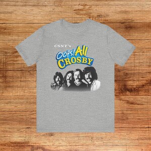 May include: A gray t-shirt featuring a graphic of four men and the text "CSNY's Oops! All Crosby" in blue and yellow. The t-shirt is made of a soft material and has a crew neck.