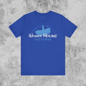 May include: Royal blue t-shirt featuring a light blue boat graphic within a semi-circle, with the text "Werner Herzog Pictures" in white and light blue. The shirt is a solid color and has short sleeves.